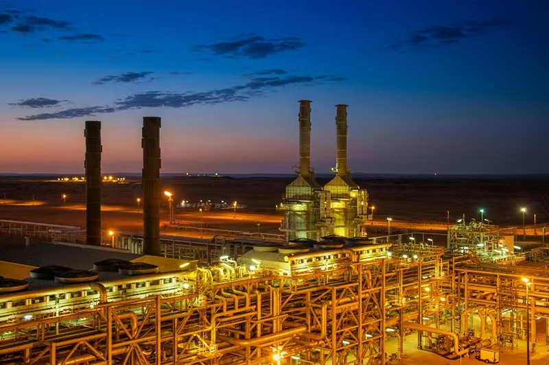 A large oil refinery at sunset, glowing with bright explosion-proof lights as the day transitions to night.