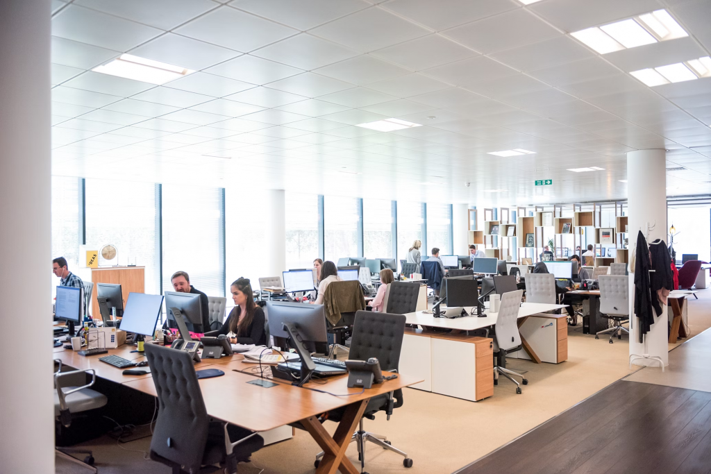 A busy office featuring individuals at desks, illuminated by overhead panel lights, engaged in their work