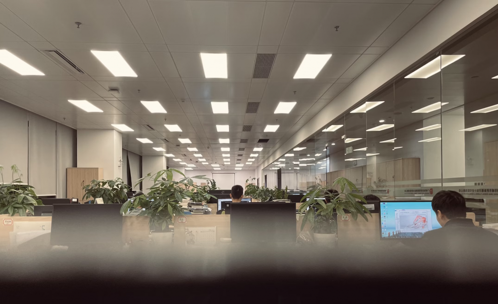 A large office space featuring employees at desks, all well lit by modern led panel lights.