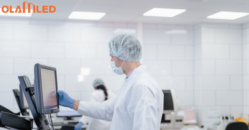 A man in a lab coat works on a computer in a cleanroom environment, illuminated by bright, sterile lighting.