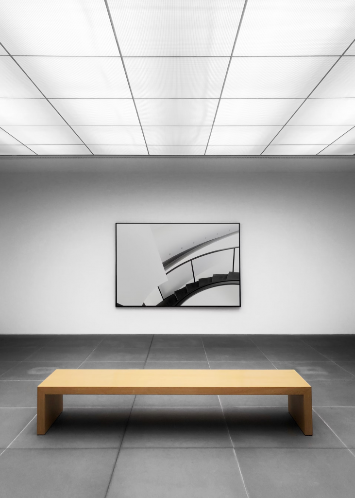 A vacant room with a bench facing a painting, softly lit by a led panel light on the ceiling.